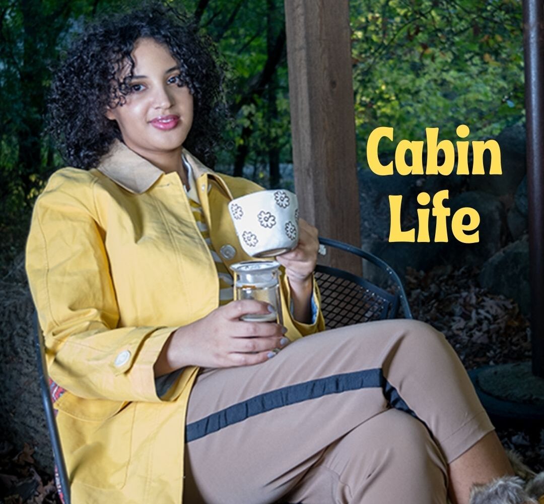 Always Uttori Cabin Life. Woman wearing yellow coat, brown pants, and holding a cup.