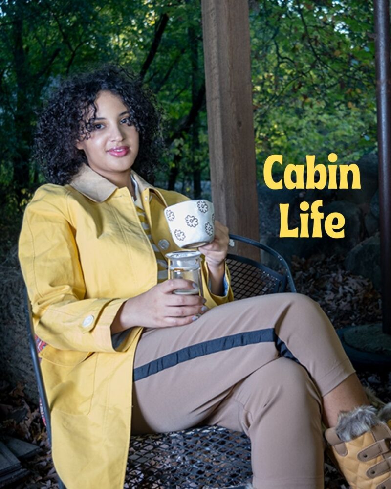Cabin Life, Always Uttori. Woman in forest wearing yellow coat, brown pants, boots and holding a cup. 